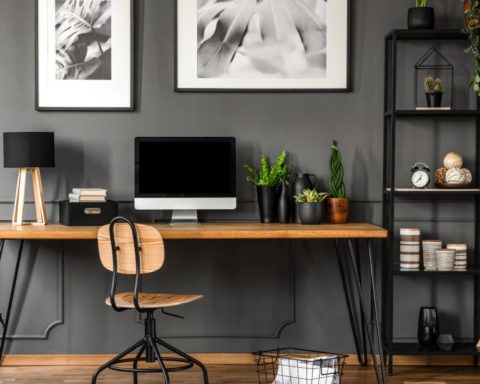 A home office with a wooden desk, adjustable chair, and computer sitting on the desk. Next to the desk is a shelving unit.
