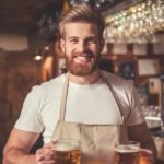 standing man with beard wearing beige apron holding two large mugs filled with golden beer inside a brewery next to bar area