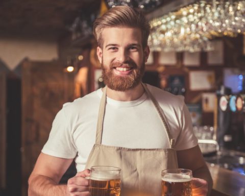 standing man with beard wearing beige apron holding two large mugs filled with golden beer inside a brewery next to bar area