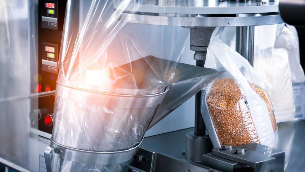 A close-up of a machine in a factory filling plastic bags with buckwheat and sealing them for distribution.
