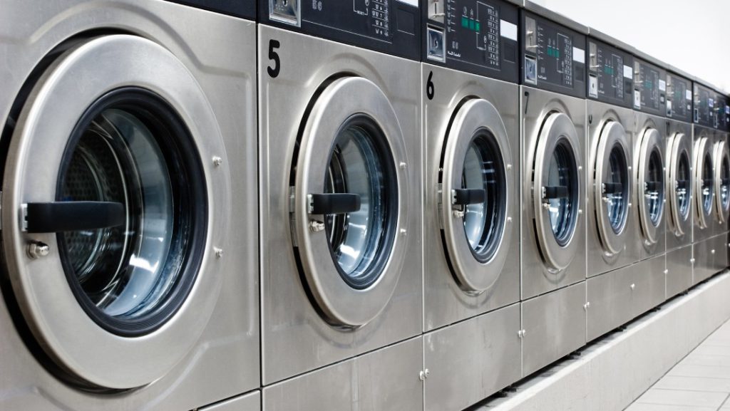 A brightly lit laundromat with a line of shiny, silver, numbered washing machines, open and ready for customers to use.