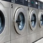 A brightly lit laundromat with a line of shiny, silver, numbered washing machines, open and ready for customers to use.