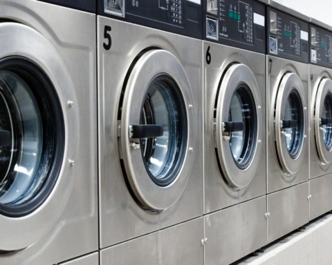 A brightly lit laundromat with a line of shiny, silver, numbered washing machines, open and ready for customers to use.