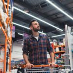 A man with a beard pushing a shopping cart down an aisle filled with tools and equipment in a hardware store.
