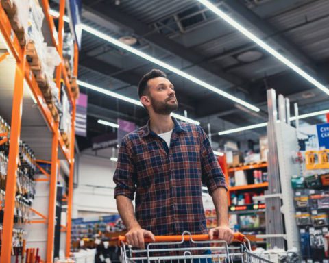 A man with a beard pushing a shopping cart down an aisle filled with tools and equipment in a hardware store.