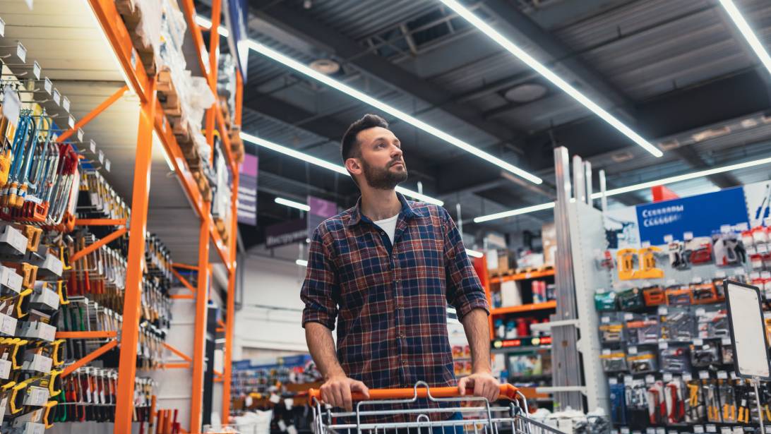 A man with a beard pushing a shopping cart down an aisle filled with tools and equipment in a hardware store.