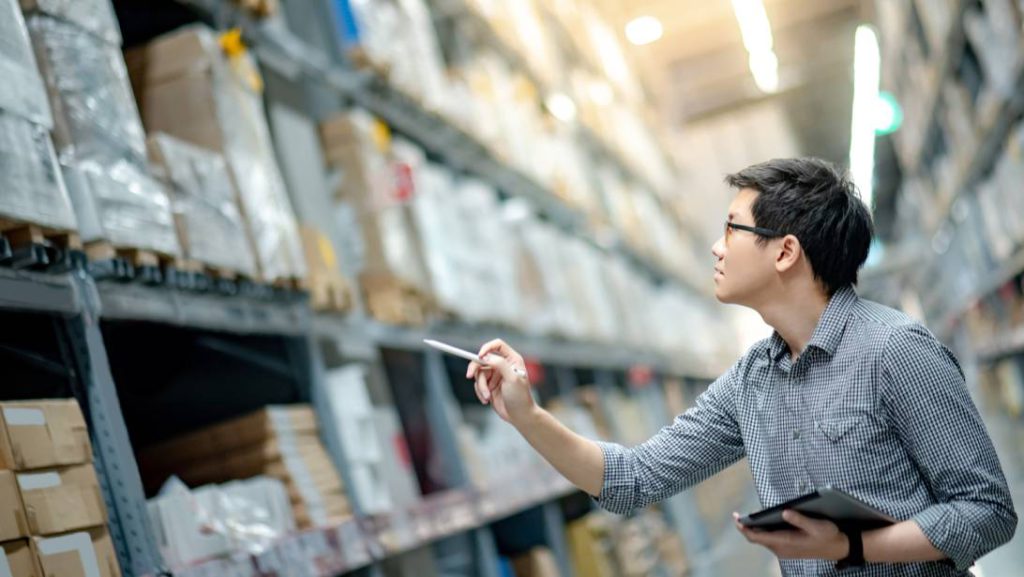 A young man with glasses holds a clipboard and looks up at shelving units in a warehouse. He holds out his pen as if counting.