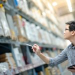 A young man with glasses holds a clipboard and looks up at shelving units in a warehouse. He holds out his pen as if counting.