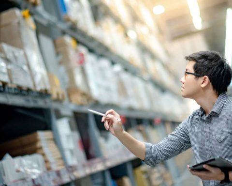 A young man with glasses holds a clipboard and looks up at shelving units in a warehouse. He holds out his pen as if counting.