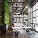 A contemporary office with ceiling to floor windows, multiple work stations, and greenery/plants everywhere.